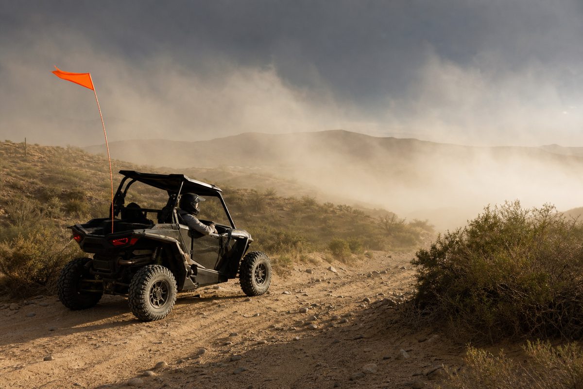 A side-by-side rider stops on a rocky desert trail as wind drives dust across the route and bends the orange whip flag
